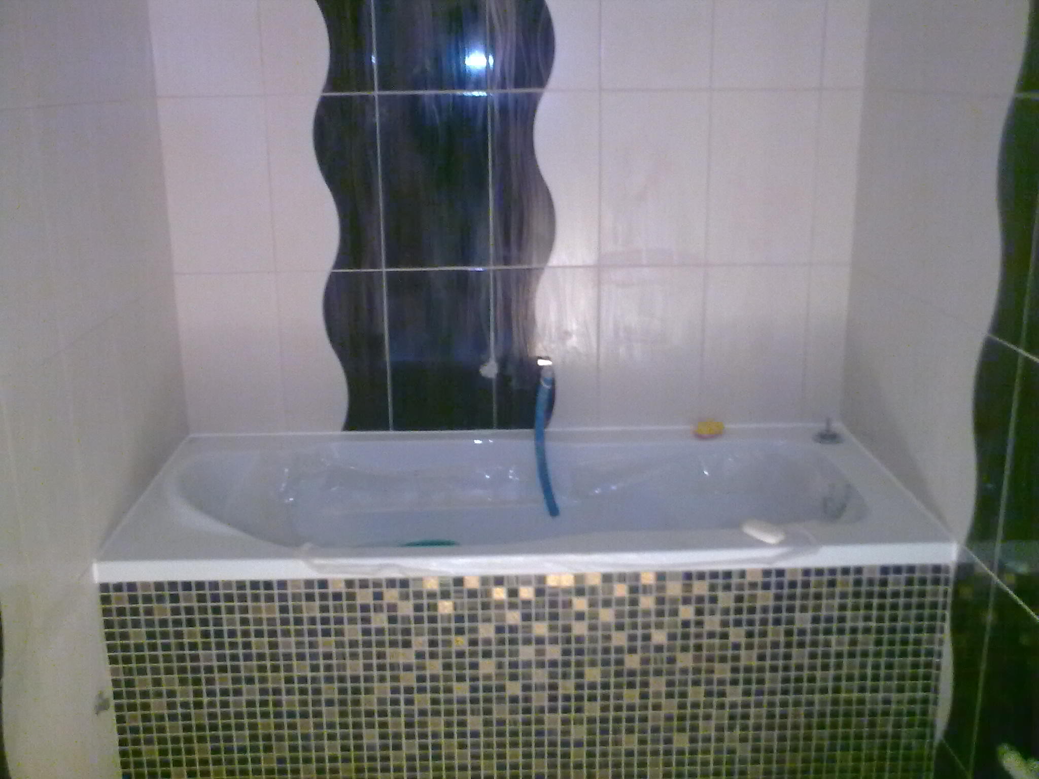 Biała wanna obudowana mozaiką w szaro-złote kwadraciki, na ścianie białe płytki z dekoracyjnym pionowym pasem z ciemnych płytek o falistym kształcie.